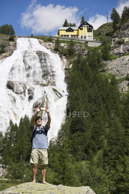 Joven tomando selfie smartphone frente a la cascada Toce, Formazza, Verbania, Piemonte, Italia - foto de stock