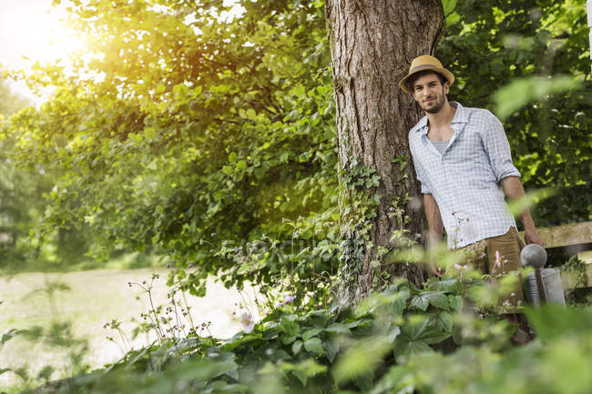 Young man leaning against tree with watering can — Stock Photo
