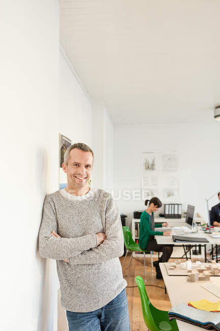 Mature man in office leaning against wall, arms crossed looking at camera smiling — Stock Photo