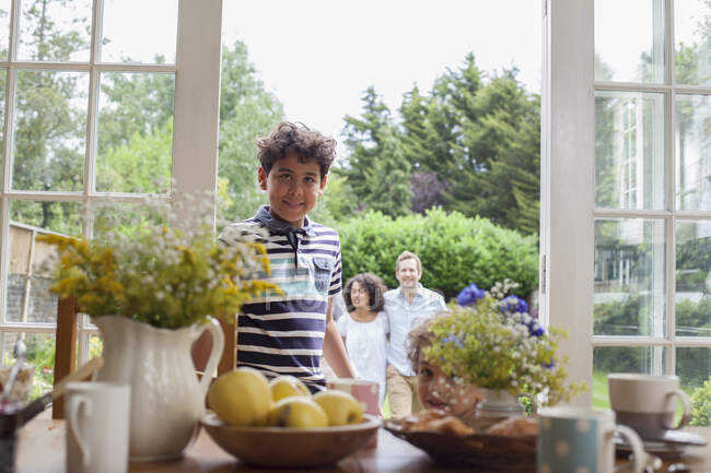 Porträt eines kleinen Jungen, der an Terrassentüren steht, Familie steht hinter ihm im Garten — Stockfoto