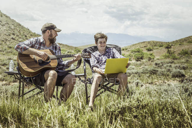 Мужчина и сын-подросток, сидящие в креслах для кемпинга, играющие на гитаре и пользующиеся ноутбуком, Бриджер, Монтана, США — стоковое фото
