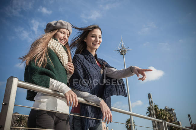 Vista de ángulo bajo de las mujeres jóvenes inclinadas contra barandilla mirando hacia otro lado señalando y sonriendo - foto de stock