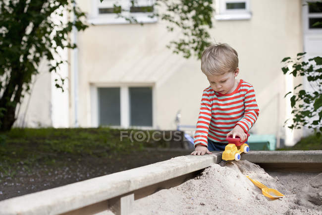 Male toddler pushing toy car in sand pit in garden — Stock Photo
