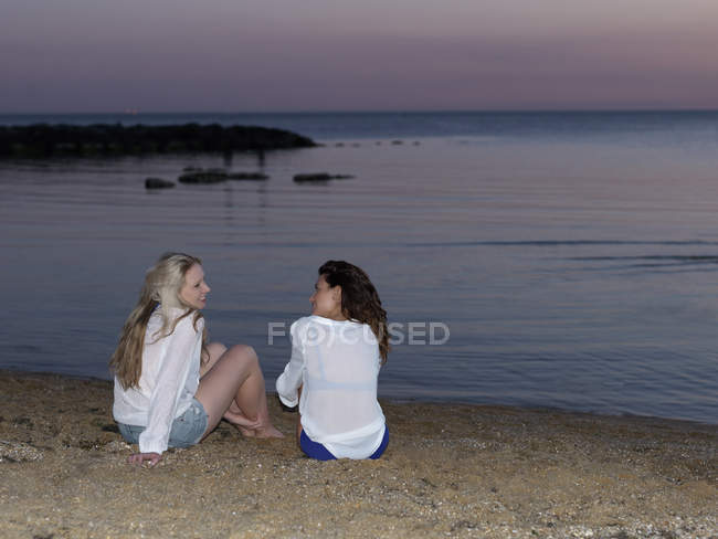 Vista trasera de dos jóvenes sentadas en la playa al atardecer - foto de stock