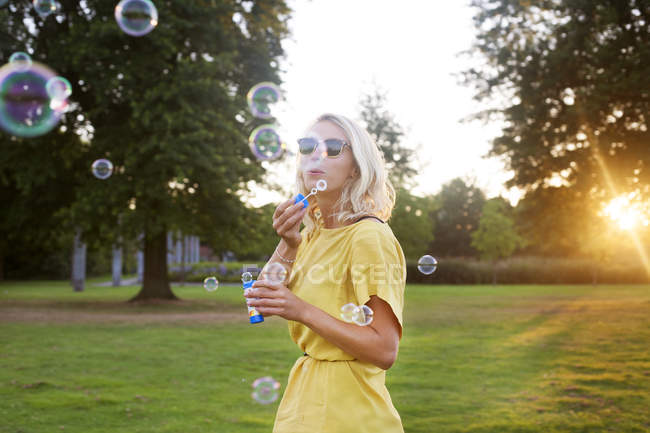 Портрет молодой женщины в жёлтом платье, дующей пузырьками в парке на закате — стоковое фото