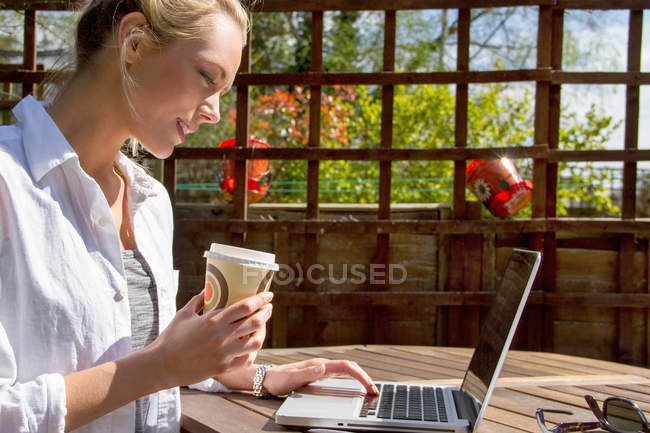Jeune femme avec café à emporter et ordinateur portable dans le jardin — Photo de stock