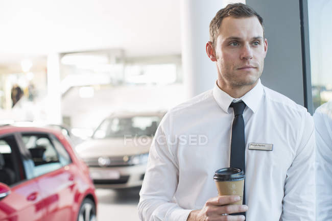 Portrait of worried salesman with takeaway coffee in car dealership — Stock Photo