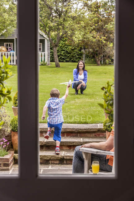 Mutter beobachtet Sohn beim Spielen im Garten mit Spielzeugflugzeug — Stockfoto