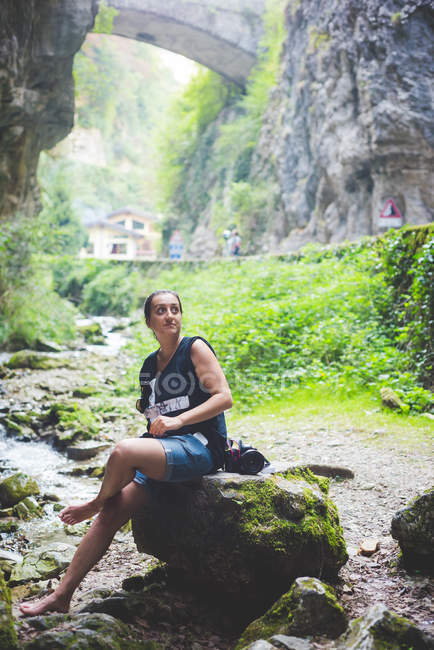 Donna seduta su roccia nel torrente, colline rocciose e ponte in pietra sullo sfondo, Garda, Italia — Foto stock