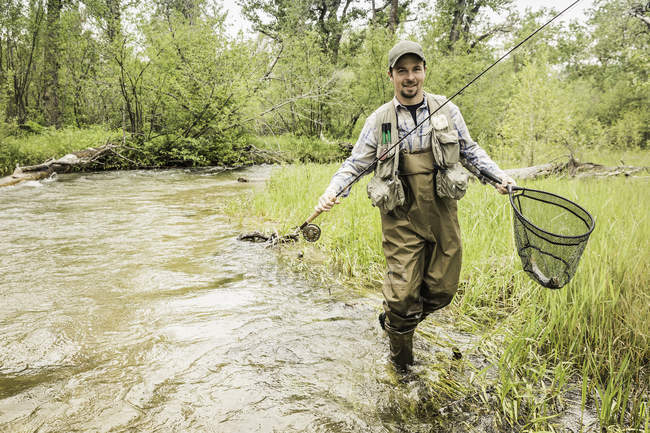 Homem no rio vestindo waders carregando rede de pesca olhando para a câmera sorrindo — Fotografia de Stock