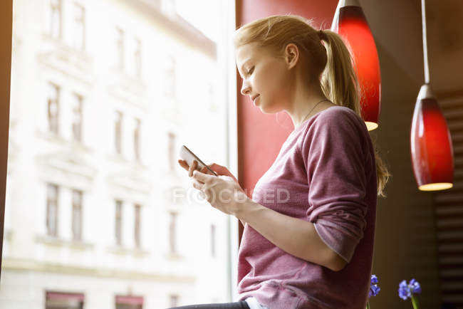 Jeune femme au café fenêtre textos sur smartphone — Photo de stock