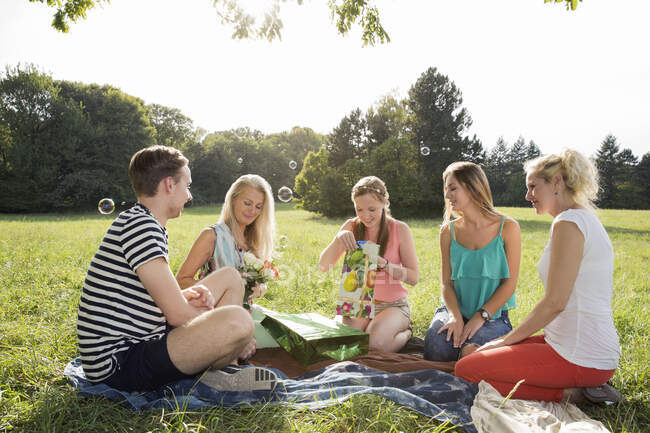 Famiglia seduta sulla coperta picnic nel parco dando donna matura fiori e regali sorridenti — Foto stock