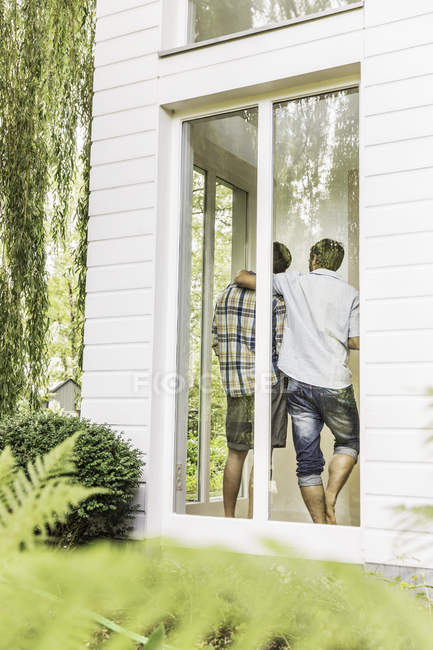 Vista trasera de dos hombres abrazando detrás de la ventana de la casa - foto de stock