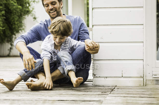 Hombre adulto medio y su hijo riendo y cosquillas pies en el porche - foto de stock