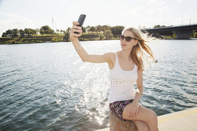 Молодая женщина, сидящая на берегу реки, делает селфи на смартфоне, остров Дунай, Вена, Австрия — стоковое фото