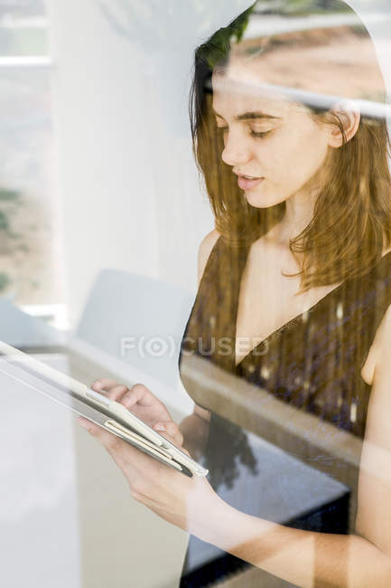 Jeune femme utilisant une tablette numérique, photographiée à travers le verre — Photo de stock