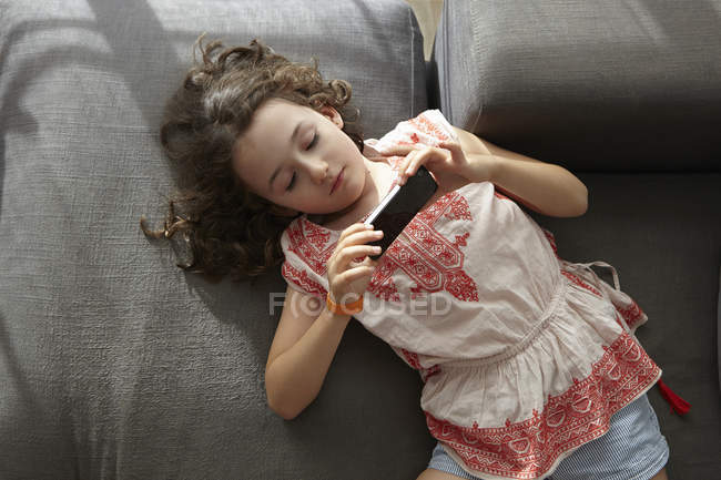Visão aérea da menina deitada no sofá da sala de estar jogando com smartphone — Fotografia de Stock