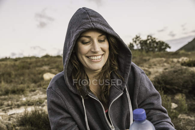 Femme portant capuche supérieure tenant bouteille d'eau regardant vers le bas souriant — Photo de stock
