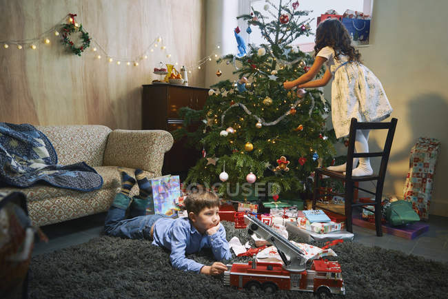 Menina organizando árvore de Natal, enquanto irmão jogando com presentes de Natal no chão da sala de estar — Fotografia de Stock