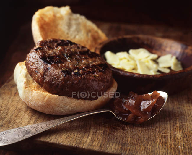 Burger in crusty bun with cheddar cheese and onion relish on wooden cutting board — Stock Photo
