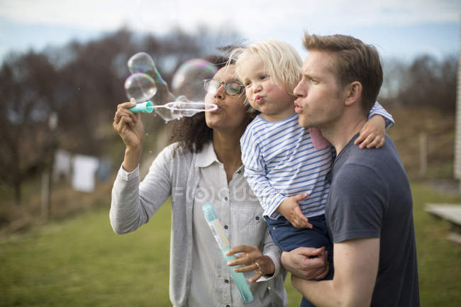 Madre soplando burbujas, padre sosteniendo hijo - foto de stock