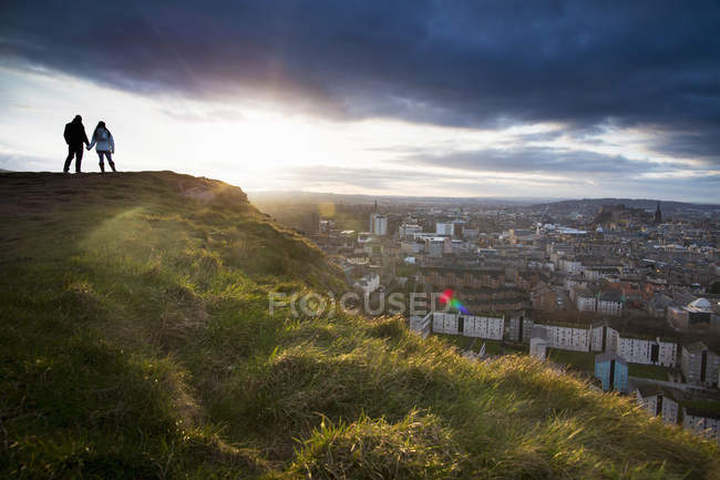 A young couple hold hands in front of the view of the City of Edinburgh from Salisbury Crags — Stock Photo
