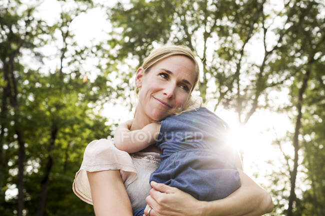 Mulher adulta média carregando filha criança no parque iluminado pelo sol — Fotografia de Stock