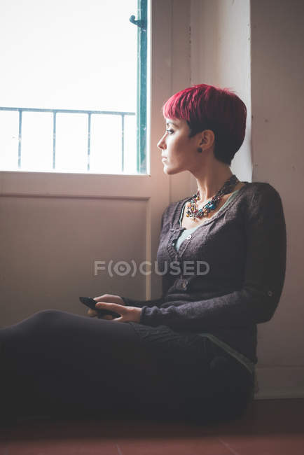 Молодая женщина сидит у окна, держа в руках смартфон — стоковое фото