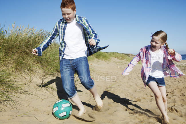 Девочка и мальчик гоняются за футболом по песку, улыбаясь — стоковое фото
