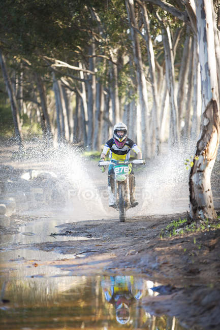 Jovem piloto de motocross masculino salpicando através da poça na floresta — Fotografia de Stock