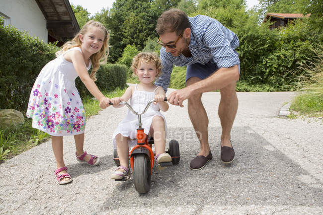 Девушка на трехколесном велосипеде с отцом и сестрой — стоковое фото