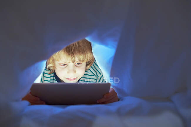 Menino sob edredão usando tablet digital — Fotografia de Stock