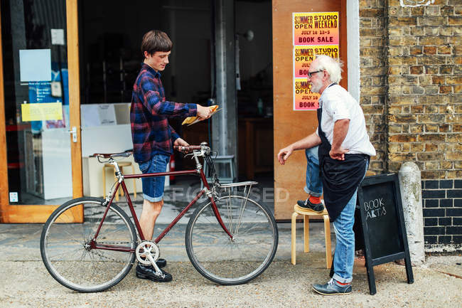 Jeune homme avec pushbike regardant le poste et debout avec l'artisan senior atelier d'impression à l'extérieur — Photo de stock