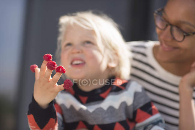 Niño con frambuesas en los dedos con la madre mirando - foto de stock