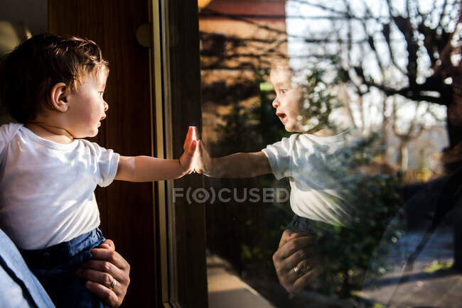 Niño en brazos del padre mirando a través de la ventana y tocando - foto de stock