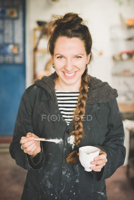 Portrait of young woman holding clay pot and paintbrush looking at camera smiling — Stock Photo