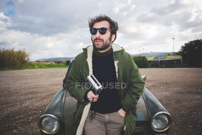 Homme adulte moyen appuyé contre la voiture vintage avec caméra de film vintage — Photo de stock