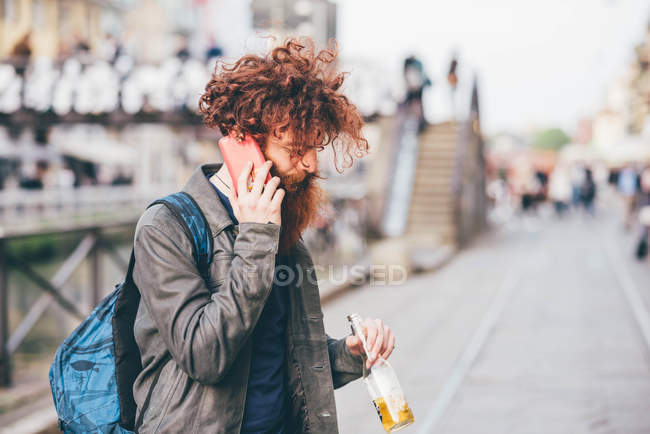Joven hipster masculino con el pelo rojo y la barba charlando en el teléfono inteligente en la calle de la ciudad - foto de stock