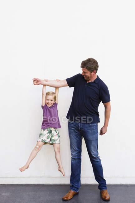Portrait of mature man with daughter hanging from his arm in front of white wall — Stock Photo
