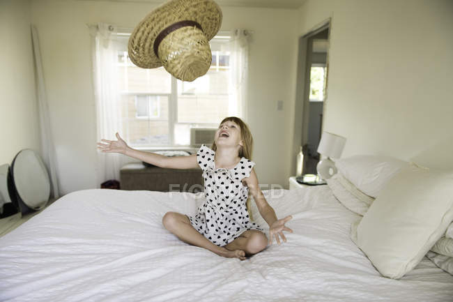 Junges Mädchen sitzt auf Bett und wirft Strohhut in die Luft — Stockfoto