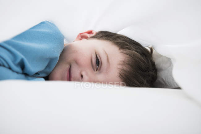 Portrait of boy lying between white bed sheets — Stock Photo