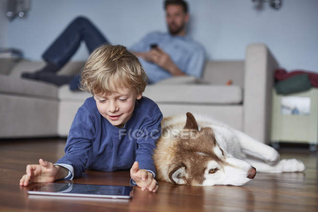 Хлопчик лежить на підлозі у вітальні з хаскі за допомогою цифрового планшета — стокове фото