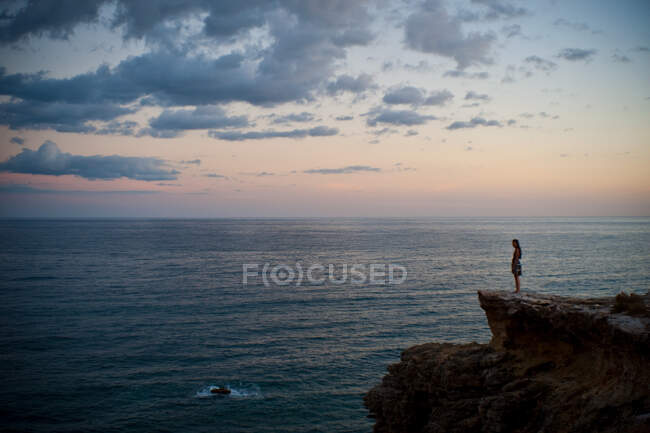 Жінка стоїть на скелі, дивлячись в море на заході сонця — стокове фото