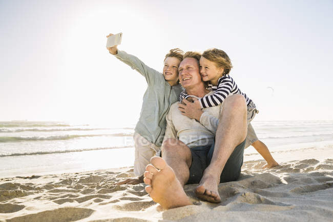 Padre e hijos en la playa usando smartphone para tomar selfie sonriendo - foto de stock