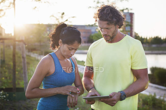 Entrenador personal masculino y mujer revisando reloj y tableta digital a orillas del río - foto de stock