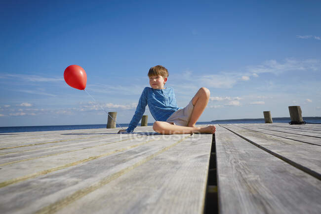 Young boy sitting on wooden pier, holding red helium balloon — Stock Photo