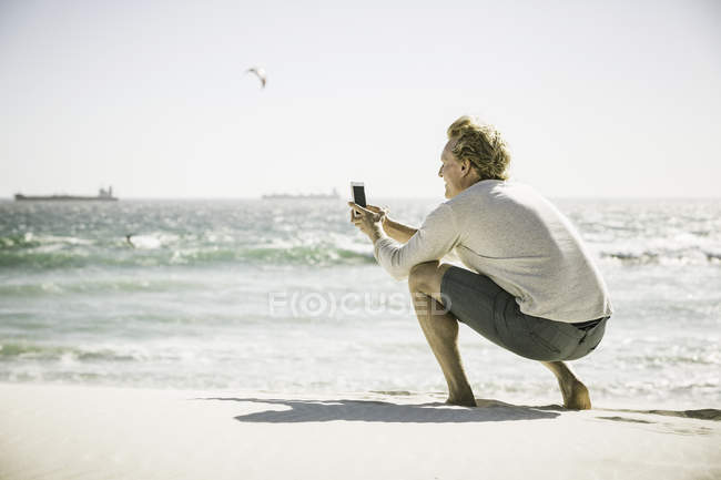 Maduro homem agachado na praia tirando fotos do mar, usando smartphone — Fotografia de Stock