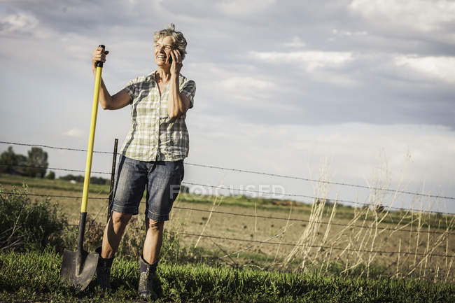 Жінка на фермі тримає лопату за допомогою смартфона, щоб зробити телефонний дзвінок — стокове фото