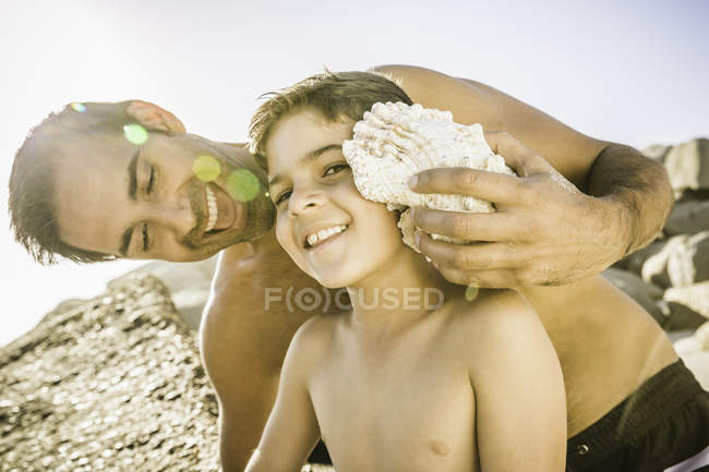 Père tenant un coquillage à l'oreille de son fils pour entendre l'océan — Photo de stock