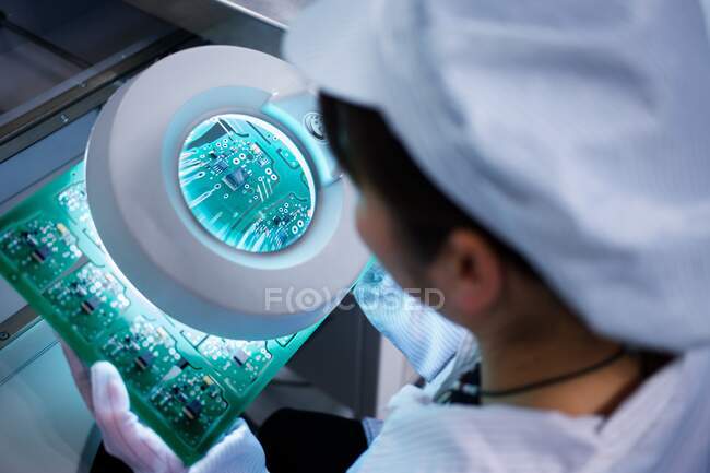 Trabajador en la fábrica de fabricación de piezas pequeñas en China mirando a través de lupa en microchips - foto de stock